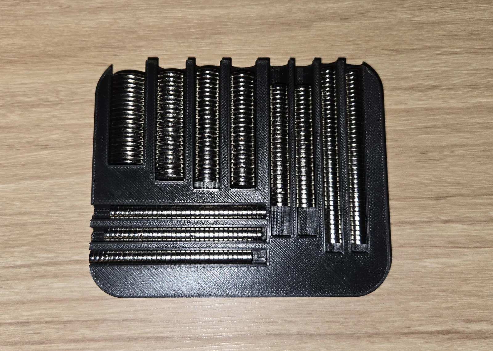 A black printed tray on a wooden bench, its nine channels filled with columns of small silver disc magnets sorted by diameter.