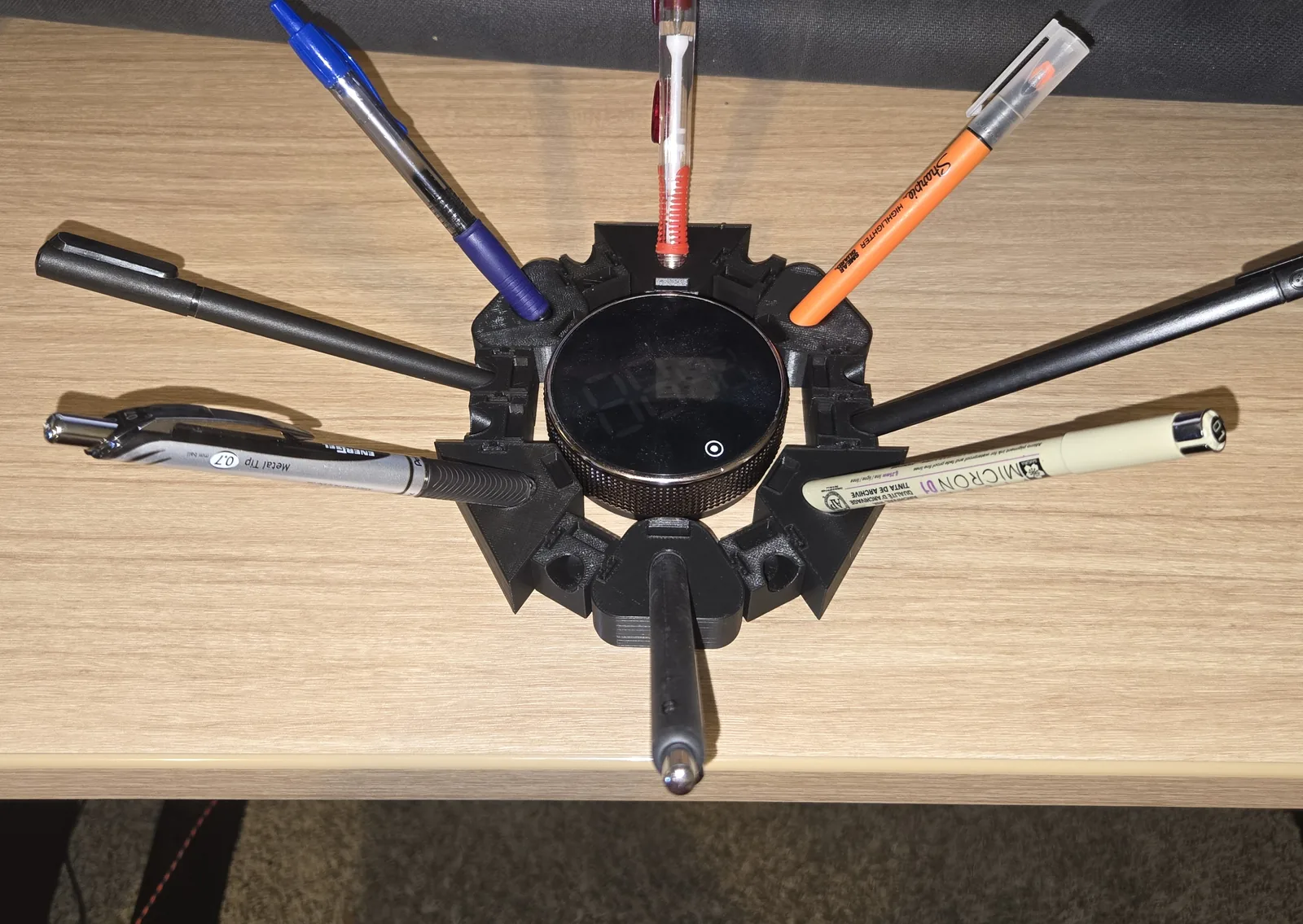 A black printed radial hub on a desk, eight pens splayed out from it in individual angled sockets, a round desk device sitting in the central well.